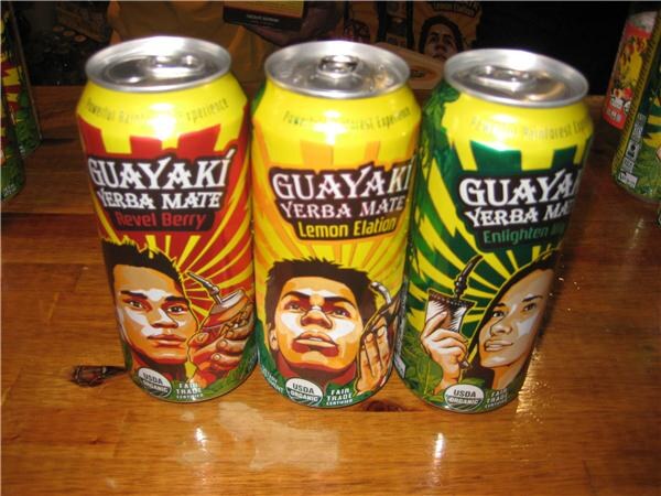 Three Guayaki Yerba Mate cans stand in a row on a wooden table: Revel Berry, Lemon Elation and Enlighten Mint, each with an illustrated face on the label.