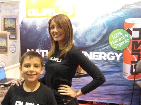 A smiling woman in a black Guru shirt stands with her hand on her hip beside a smiling boy in a black Guru shirt, in front of a wave-themed Guru banner.
