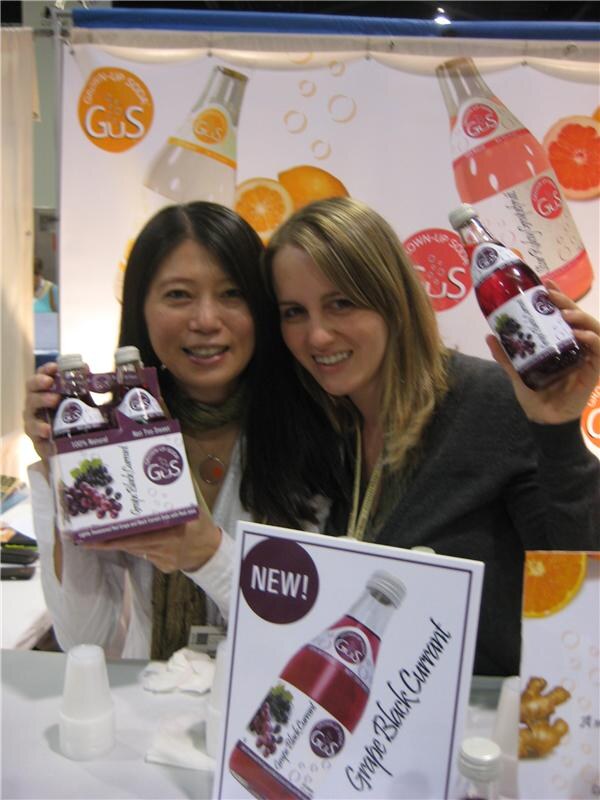 Two smiling women hold GuS Grown-Up Soda bottles, one a four-pack and one a single bottle of Grape Black Currant, in front of GuS banners with a NEW sign below.