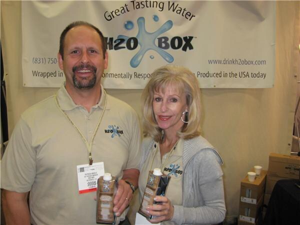A smiling man and woman in tan H2O Box shirts each hold a carton of H2O Box water in front of an H2O Box banner reading Great Tasting Water.