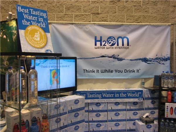 An H2OM booth with a banner reading Think It While You Drink It, a Best Tasting Water in the World sign, a screen, bottles on glass shelves and stacked cases.
