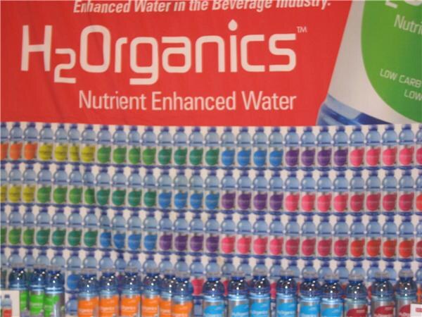 A red H2Organics Nutrient Enhanced Water banner above a wall of stacked water bottles with multicolored labels in yellow, green, blue, purple, pink and orange.