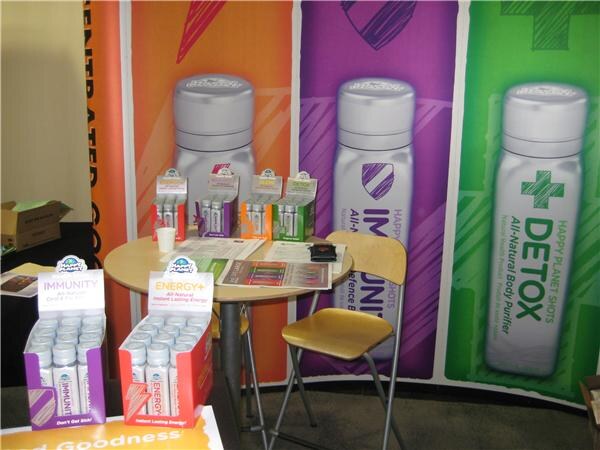 A small round table holds shot boxes in front of large Happy Planet Shots banners for Immunity, Energy and Detox. Display boxes of Immunity and Energy+ shots sit in the foreground.