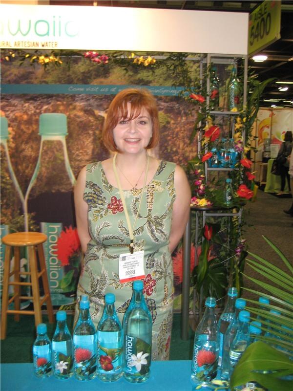 A smiling woman with red hair and a badge stands at a booth with Hawaiian Springs water bottles on the table in front of her, tropical flowers and a large bottle backdrop behind.