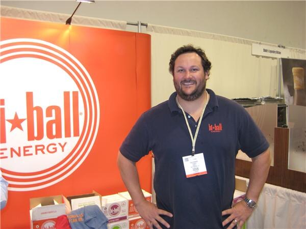 A smiling bearded man in a navy Hi-Ball polo with a badge stands with hands on hips in front of an orange Hi-Ball Energy banner, with cases of product at the bottom.