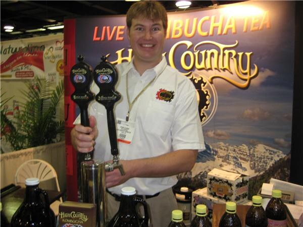 A smiling man in a white shirt holds a black draft tap handle at a booth with a Live Kombucha Tea High Country banner and growlers and bottles on the counter.