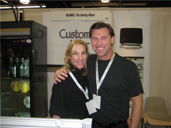 A smiling woman with blonde hair and a smiling man in black, both wearing lanyards, pose with arms around each other at a booth with a Hijinks: The Energy Mixer sign and a refrigerated case.