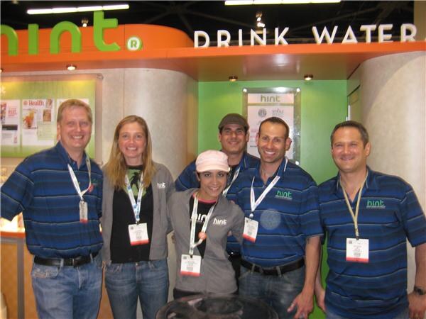 Five people in Hint shirts and badges pose together in front of the Hint Drink Water booth, which has a green and orange sign and wall panels.