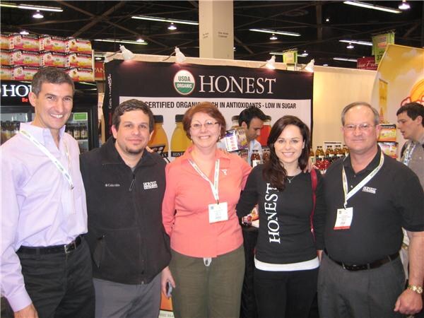 Five smiling people with show badges pose in front of an Honest Tea banner reading "Honest" and "Certified organic"; one wears a Columbia fleece, and another person stands behind them.