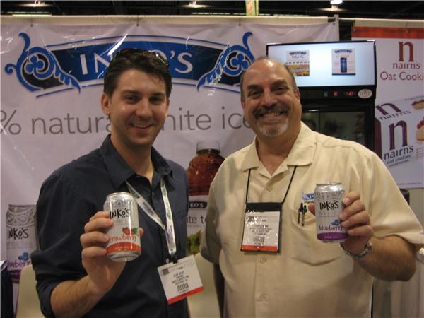 Two smiling men with show badges hold up Inko's white tea cans, strawberry and blueberry, in front of an Inko's banner, with a Nairns oat cookies sign at right.