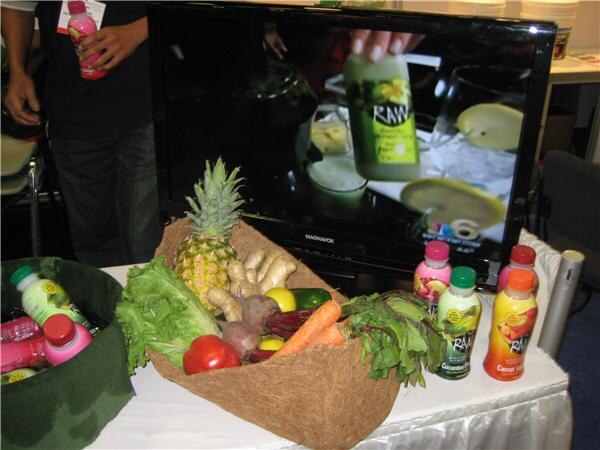Table with a basket of fresh produce including pineapple, lettuce, carrots and tomatoes beside RAAW juice bottles, with a Magnavox screen behind them.