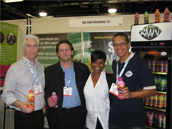 Three men and a woman pose in a booth labeled Raw Foods International, LLC, holding RAAW juice bottles, with a lit RAAW refrigerator and a Ceiflore banner at left.