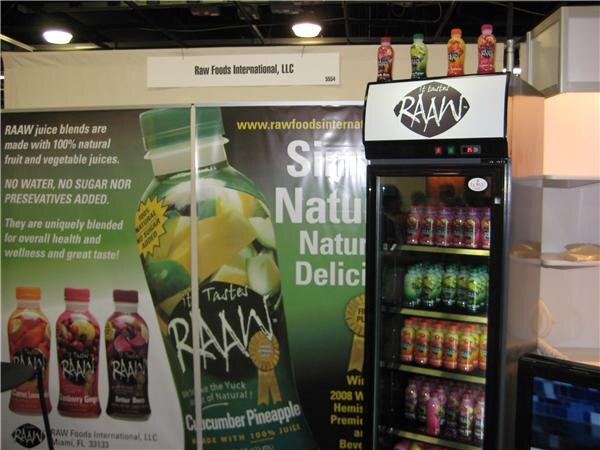 Booth for Raw Foods International, LLC with a large Cucumber Pineapple RAAW bottle graphic, juice bottles on the wall, and a lit refrigerator stocked with RAAW juices.