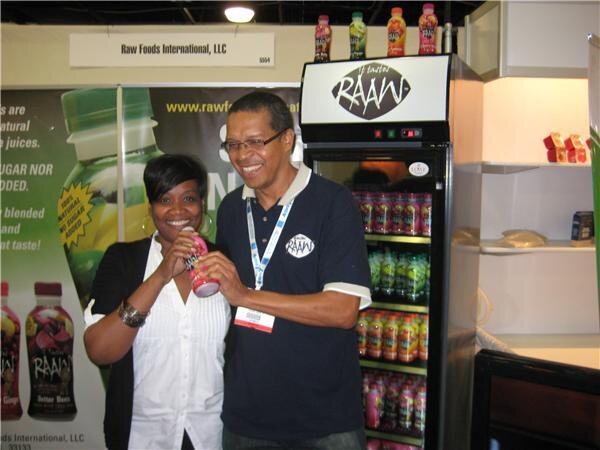 A smiling woman and a man in a navy polo hold a pink RAAW juice bottle beside a RAAW refrigerator in the Raw Foods International, LLC booth.