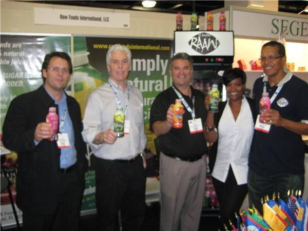 Five people, four men and one woman, hold RAAW juice bottles in the Raw Foods International, LLC booth, with a RAAW refrigerator and small flags nearby.