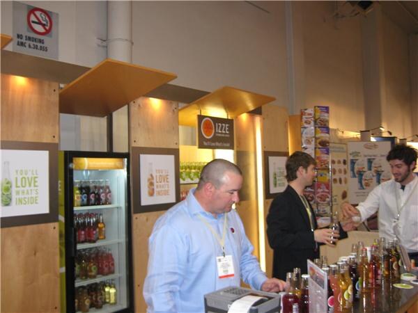 Izze booth with wood-paneled walls and "You'll love what's inside" signs, a refrigerator of bottled drinks, and three men behind the counter, one using a register.
