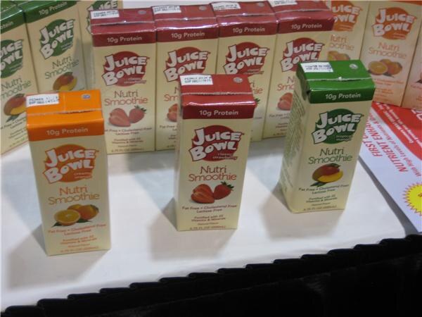 Juice Bowl Nutri Smoothie cartons stand on a white table in orange, red and green, with more cartons stacked behind. Each carton is labeled 10g Protein.