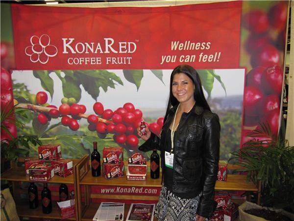 A smiling woman with long dark hair and a black leather jacket holds a small bottle in front of the KonaRed Coffee Fruit booth, with boxes and bottles on wooden shelves behind her.