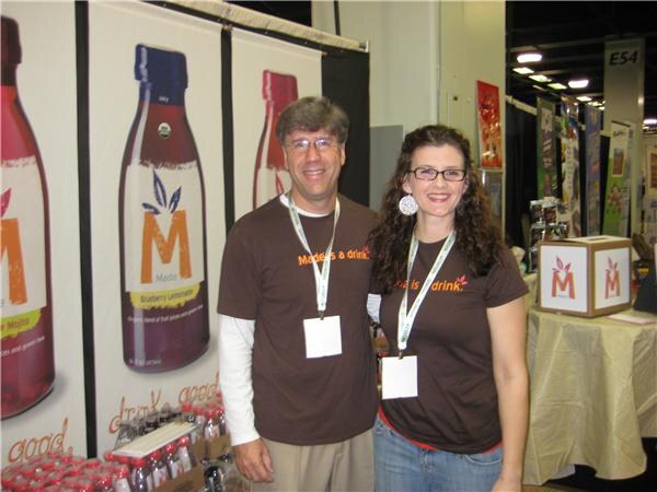 A man with glasses and a woman with curly dark hair and glasses, both in brown Made t-shirts and badges, stand together in front of large Made bottle banners.