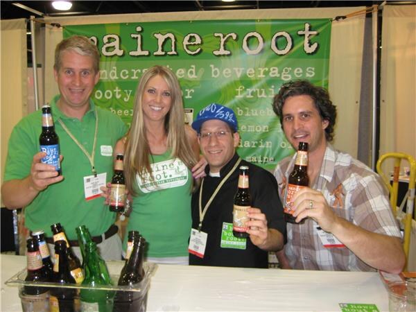 Four people hold bottles of Maine Root soda behind a counter in front of a green Maine Root banner. Several bottles sit in a tray of ice on the counter.