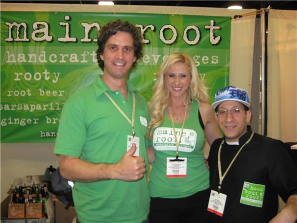 A man in a green shirt giving a thumbs up, a blonde woman in a green Maine Root top and a man in a blue cap and black shirt stand in front of a green Maine Root banner.