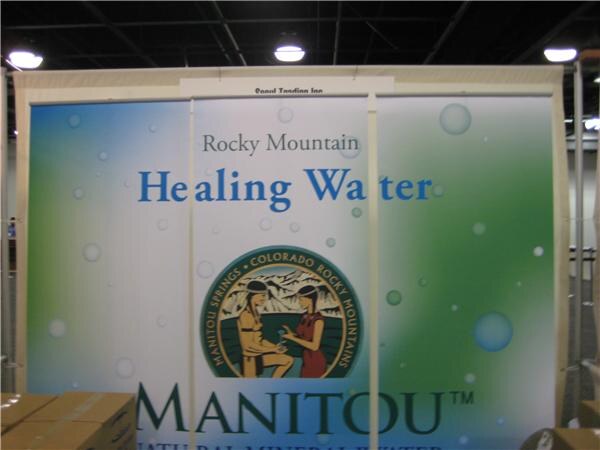 A booth backdrop reads Rocky Mountain Healing Water above the Manitou Springs Colorado Rocky Mountains logo, with cardboard boxes at the lower left.
