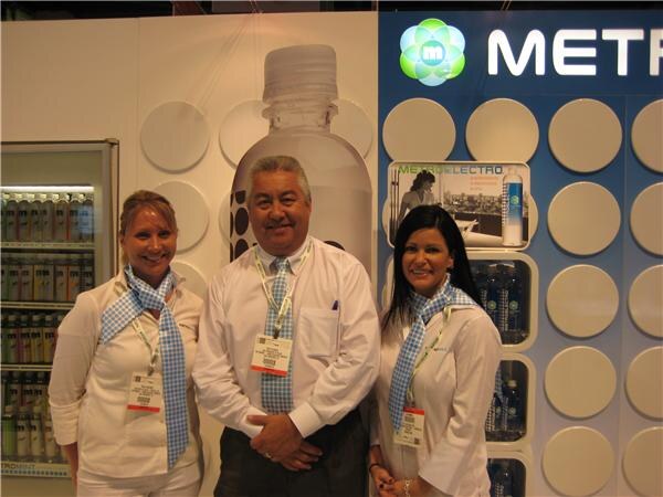 Two women and a man in white shirts and blue checkered scarves stand in front of a Metromint display with a large bottle graphic and a refrigerated cooler of bottled drinks.