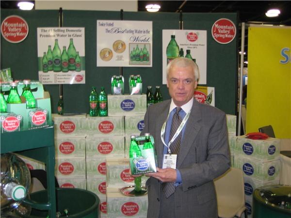 A man in a gray suit and tie holds a four-pack of green glass Mountain Valley Spring Water bottles in front of stacked cases and signs at the booth.