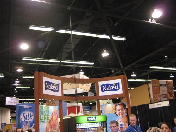 Overhead view of a booth with Naked signs above a Naked juice cooler and a Silk carton graphic at left. A few people stand below, with lights and rafters on the ceiling.