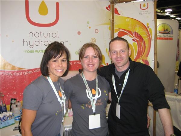 Two women and a man pose together in front of a Natural Hydration banner; the women wear gray Natural Hydration shirts and all three wear lanyards.