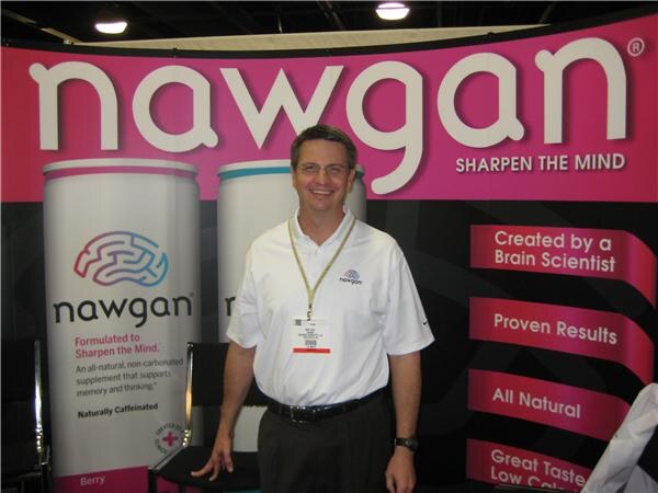 A smiling man with glasses in a white polo with a badge stands in front of a pink nawgan banner reading Sharpen the Mind, beside tall nawgan bottle graphics.