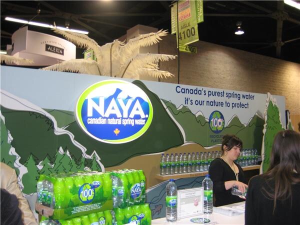 NAYA booth with a large oval NAYA Canadian natural spring water logo, a mountain mural, stacked water bottle packs, and a woman behind a counter with bottles on a shelf.