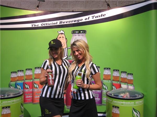 Two smiling women in referee-style striped shirts hold New Leaf bottles in front of a green backdrop reading "The Official Beverage of Taste" with rows of bottles pictured.