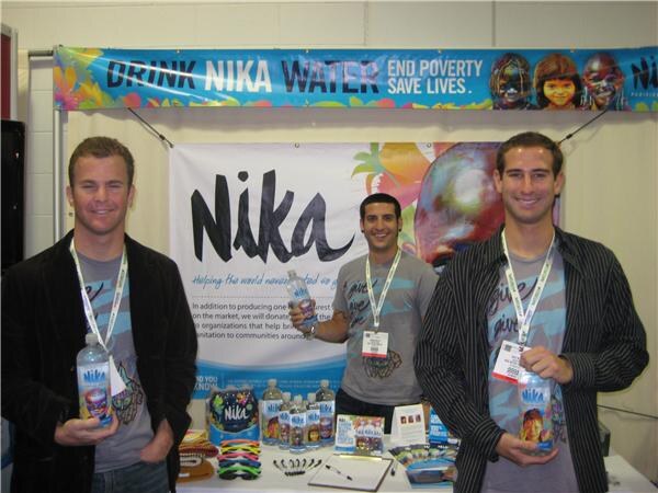 Three smiling men with lanyards hold Nika Water bottles behind a table, with a banner reading "Drink Nika Water, End Poverty, Save Lives" above and a Nika sign behind them.
