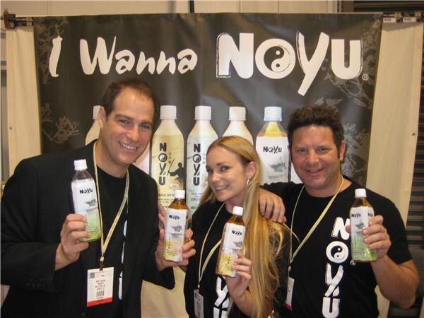 Two men and a woman with long blonde hair pose smiling, each holding a NoYu tea bottle, in front of a banner reading "I Wanna NoYu" with bottles pictured.