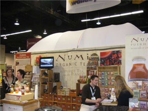 Numi Organic Tea booth with a large white canopy, tea boxes on wooden shelves, and a monitor. A man with a lanyard stands at the counter near a woman, and two women stand at left.
