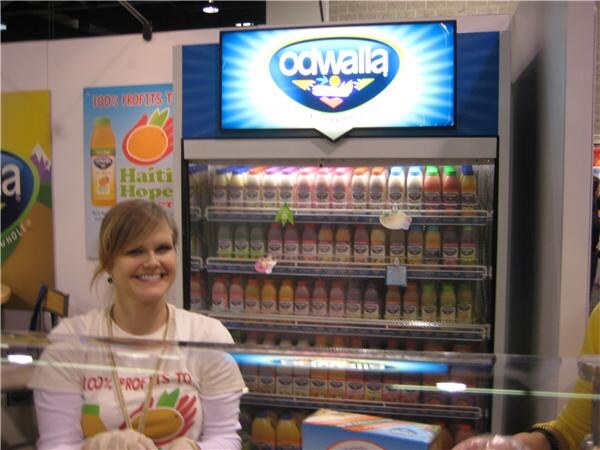 A smiling woman in a white shirt stands behind a counter in front of an Odwalla refrigerated case filled with juice bottles, under a lit Odwalla sign.