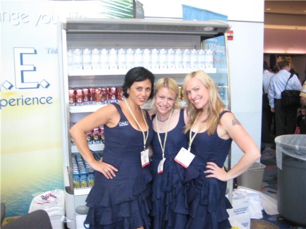 Three smiling women in navy ruffled dresses and lanyards pose with hands on hips in front of an open refrigerated display of O.N.E. bottled drinks.