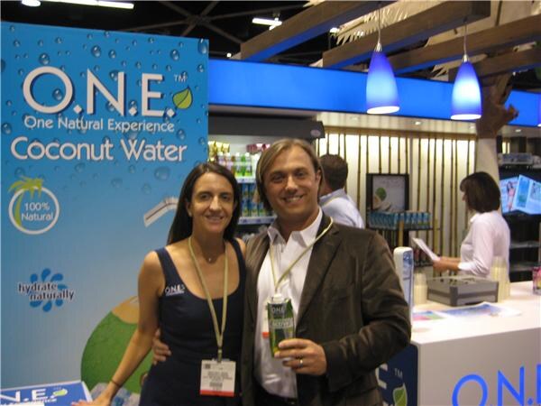 A smiling woman and man with lanyards stand beside a blue O.N.E. Coconut Water banner. The man holds a carton of the drink, and a booth counter is behind them.
