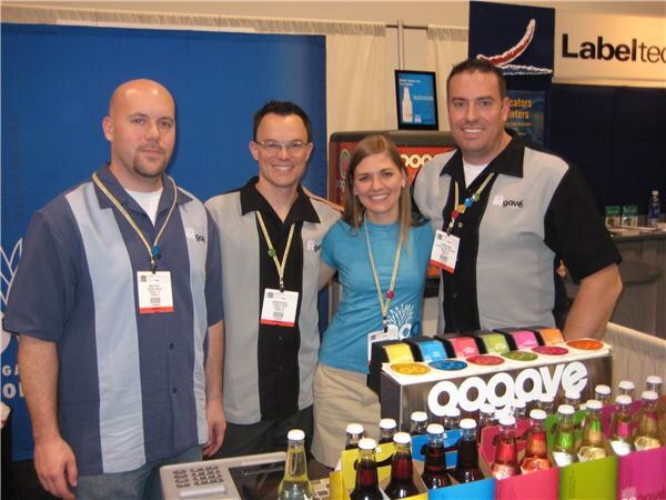 Three men and a woman with lanyards smile behind a counter of Oogave bottled sodas, with a Labeltec sign visible in the background.