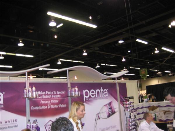 Penta booth with a purple banner about patented water, bottles on shelves, a blonde woman in white in front, and other people near a counter.