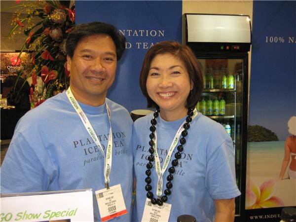 A smiling man and woman in light blue Plantation Iced Tea T-shirts and show lanyards, the woman wearing a black bead lei, in front of a blue banner and a drink cooler.