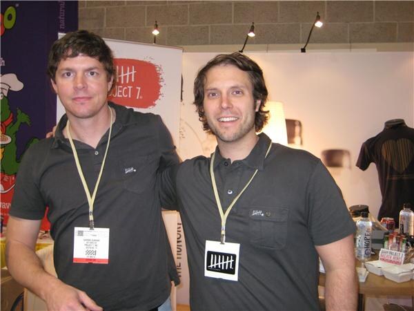Two smiling men in dark gray polo shirts and lanyards stand side by side in front of a Project 7 banner, with bottles and a shirt on display behind them.