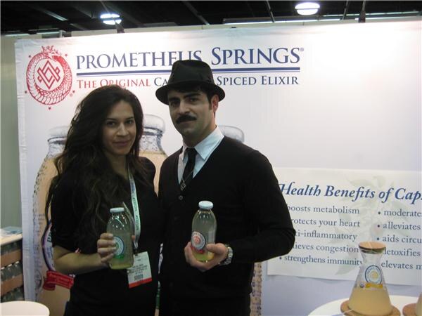 A woman and a man in a black hat and mustache each hold a small bottle in front of a Prometheus Springs banner for a capsaicin spiced elixir.