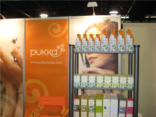 Pukka booth with an orange banner, an orange chair, and shelves holding bottles and boxes of tea in front of a photo of a woman's face.