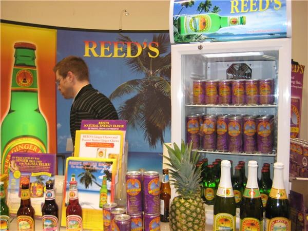 Reed's booth with a large green bottle graphic, a refrigerated cooler of purple cans, bottles, a pineapple, brochures, and a man in a striped shirt looking away.