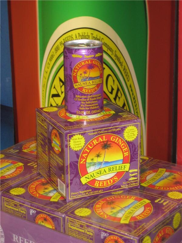 A purple can of Reed's Natural Ginger Nausea Relief sits on a four-can box, stacked on more boxes in front of a green and orange Reed's display.