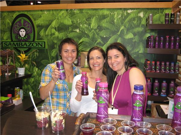 Three smiling women at the Sambazon booth, two holding purple Sambazon bottles and one a can, with a jungle backdrop, shelves of bottles and sample cups on the counter.