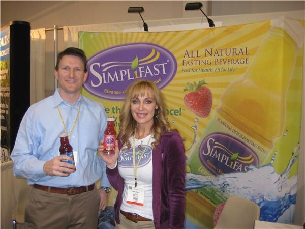 A man in a light blue shirt and a woman with long blonde hair in a purple jacket each hold a bottle of SimpliFast beverage in front of a yellow SimpliFast banner with a strawberry.