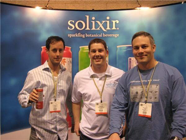 Three smiling men wearing lanyards and badges stand in front of a blue Solixir sparkling botanical beverage backdrop; the man on the left holds a red can.